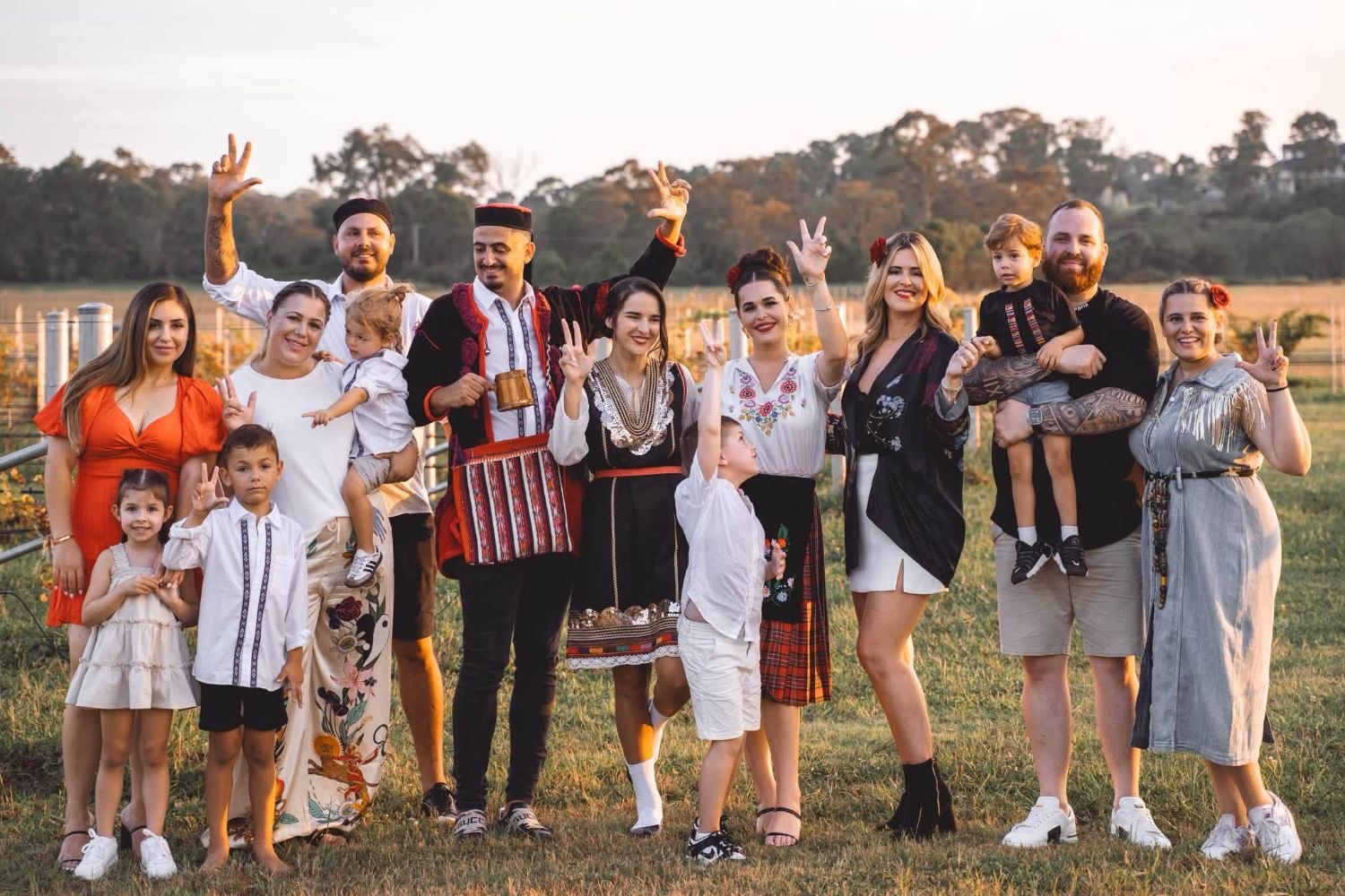 A big family group, some in traditional dress, laughing and waving together at sunset in a vineyard