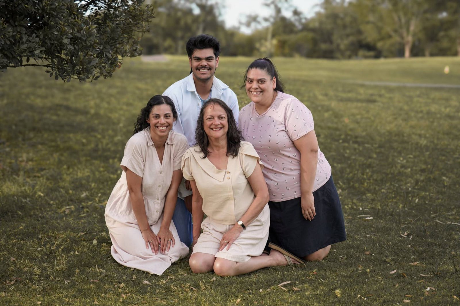 A mum and her three grown-up children smiling together on the grass in a park